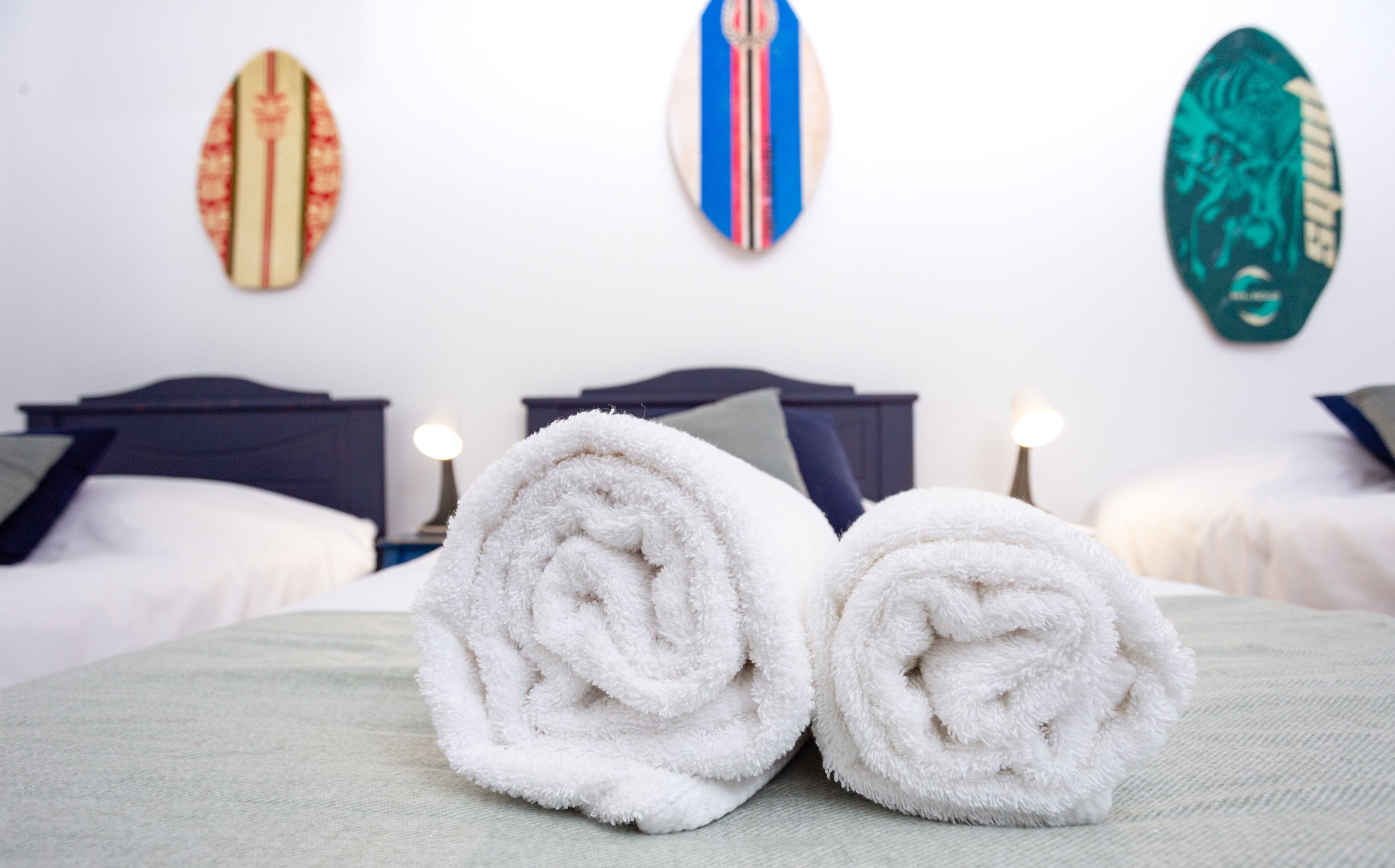 A neatly made bed with two rolled white towels, a dark headboard, and three surfboards on the wall.