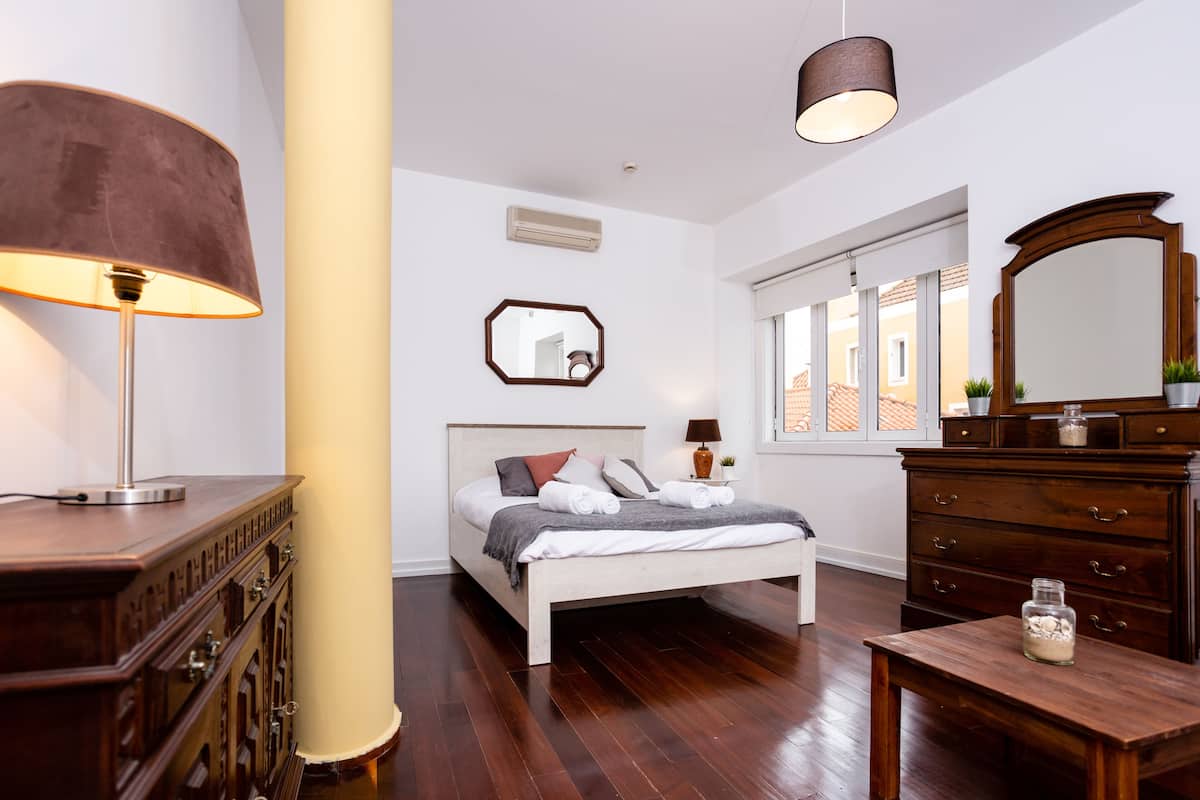 A bedroom with a bed, bedside tables, a dresser, a lamp, a mirror, and a window with a view of buildings.