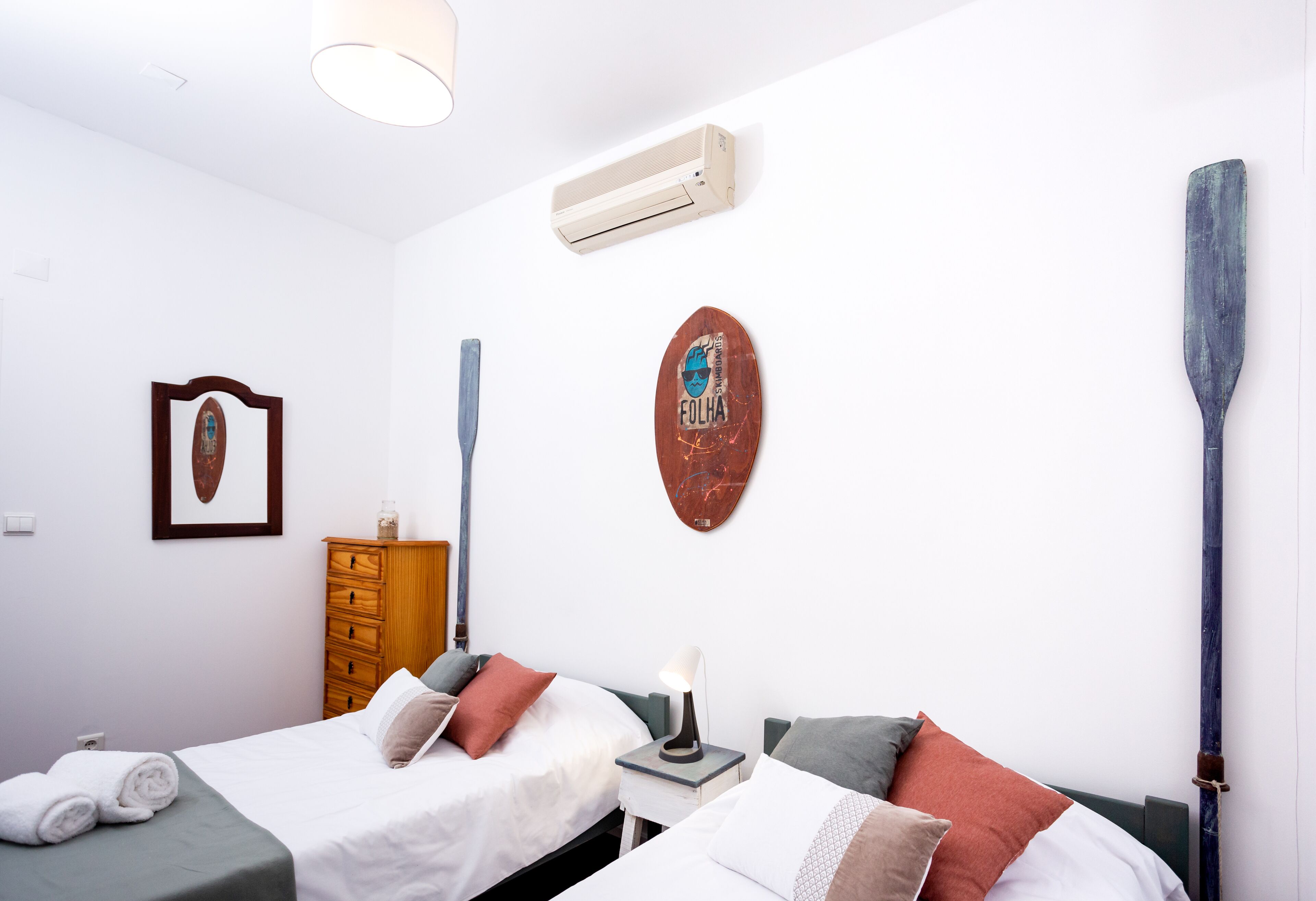 A bedroom with two beds, a wooden dresser, a wall-mounted air conditioner, and a surfboard decoration.