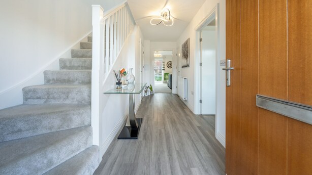 Un couloir moderne avec un escalier recouvert de moquette, une table en verre agrémentée d’objets décoratifs et une porte en bois.