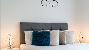 Un lit bien fait, doté d’une tête de lit rembourrée de couleur grise, de draps blancs et de trois coussins décoratifs.