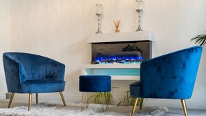Une pièce avec une cheminée, deux fauteuils en velours bleu, un pouf assorti et un tapis blanc.