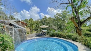 A circular pool with clear water, surrounded by lush greenery and a stone waterfall feature.