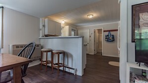 Un vaste espace de vie comprenant une table à manger en bois, deux tabourets de bar, un coin cuisine avec des meubles blancs et un téléviseur posé sur un meuble.