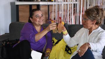 Two people sitting on a couch, holding drinks, with a coffee machine and a bowl of fruit on a table in the background.