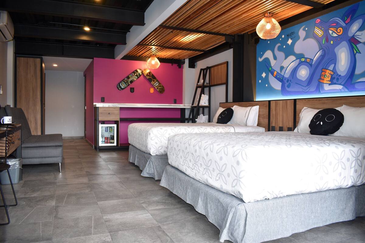 A modern hotel room with two beds, a small kitchenette, a pink accent wall, and a mural.