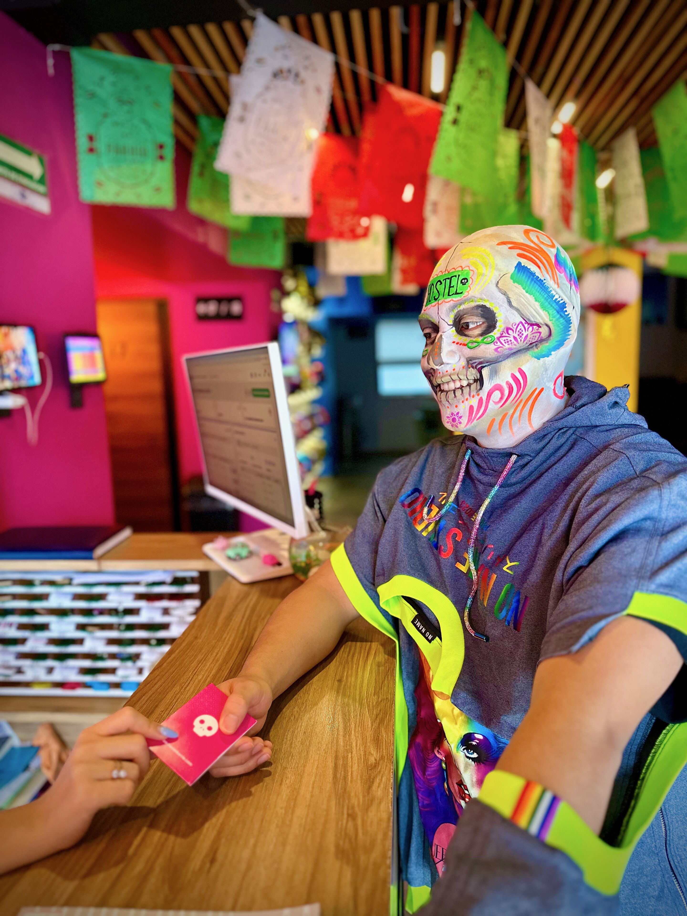 A person seated at a wooden table with a computer monitor, surrounded by colorful decorations and a vibrant pink wall.