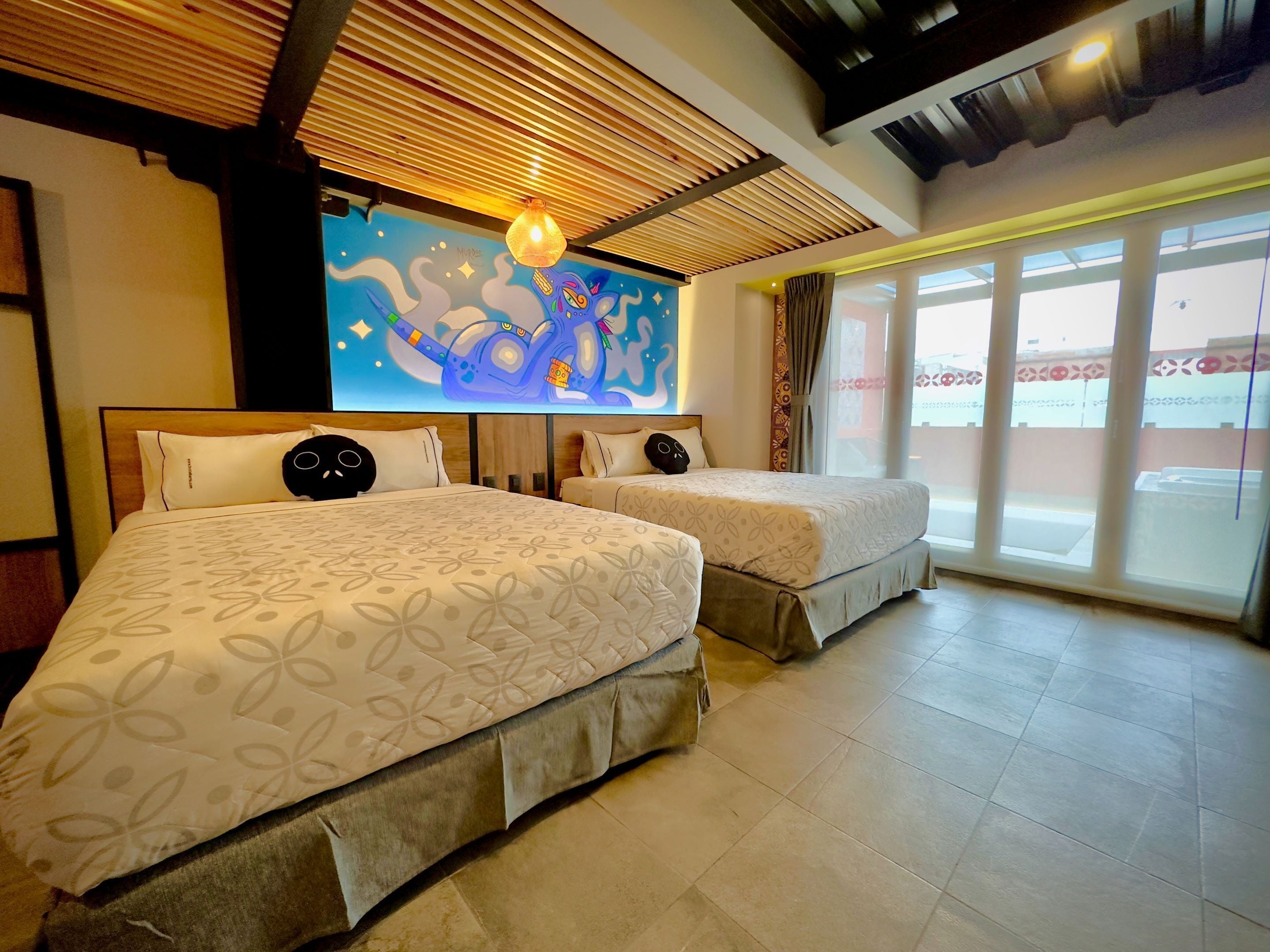 A hotel room with two beds, a large mural on the wall, and a view through the window.