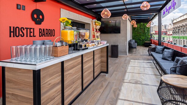 Un hostal con una pared roja, una zona de bar con diversas opciones de comida y bebida, y un área de estar con vistas a la ciudad.