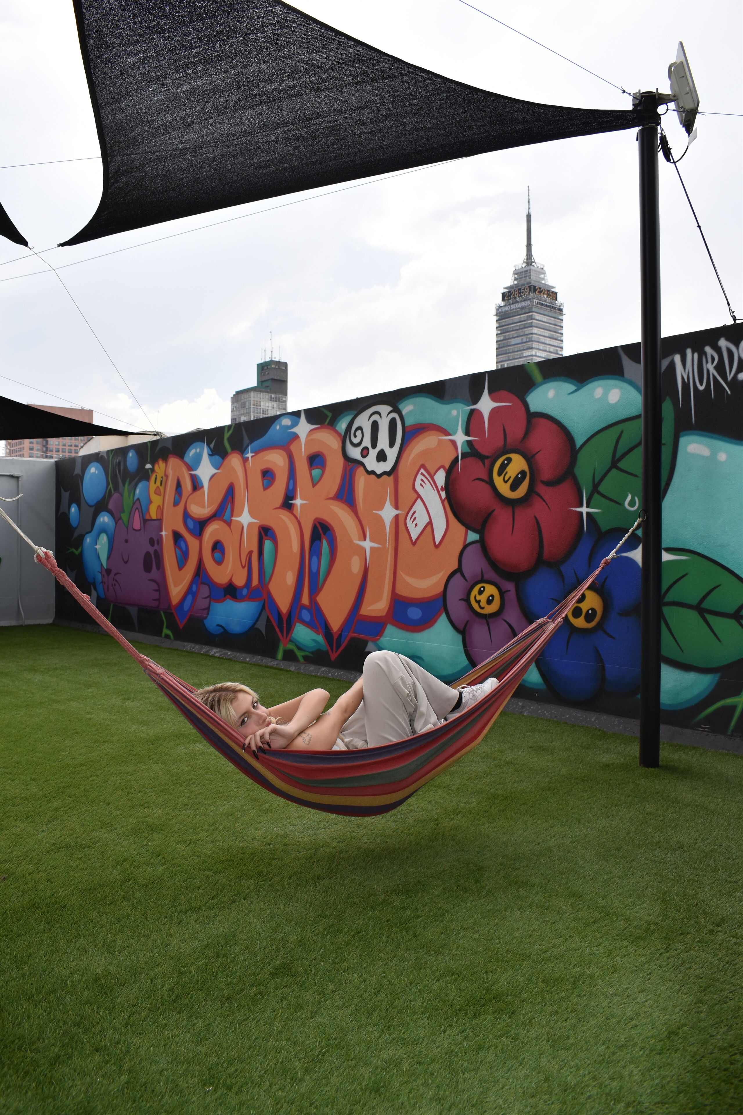 Uma pessoa relaxando em uma rede sob uma lona de sombra, com um muro de grafite colorido e um horizonte urbano ao fundo.