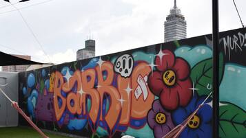 Uma pessoa relaxando em uma rede sob uma lona de sombra, com um muro de grafite colorido e um horizonte urbano ao fundo.