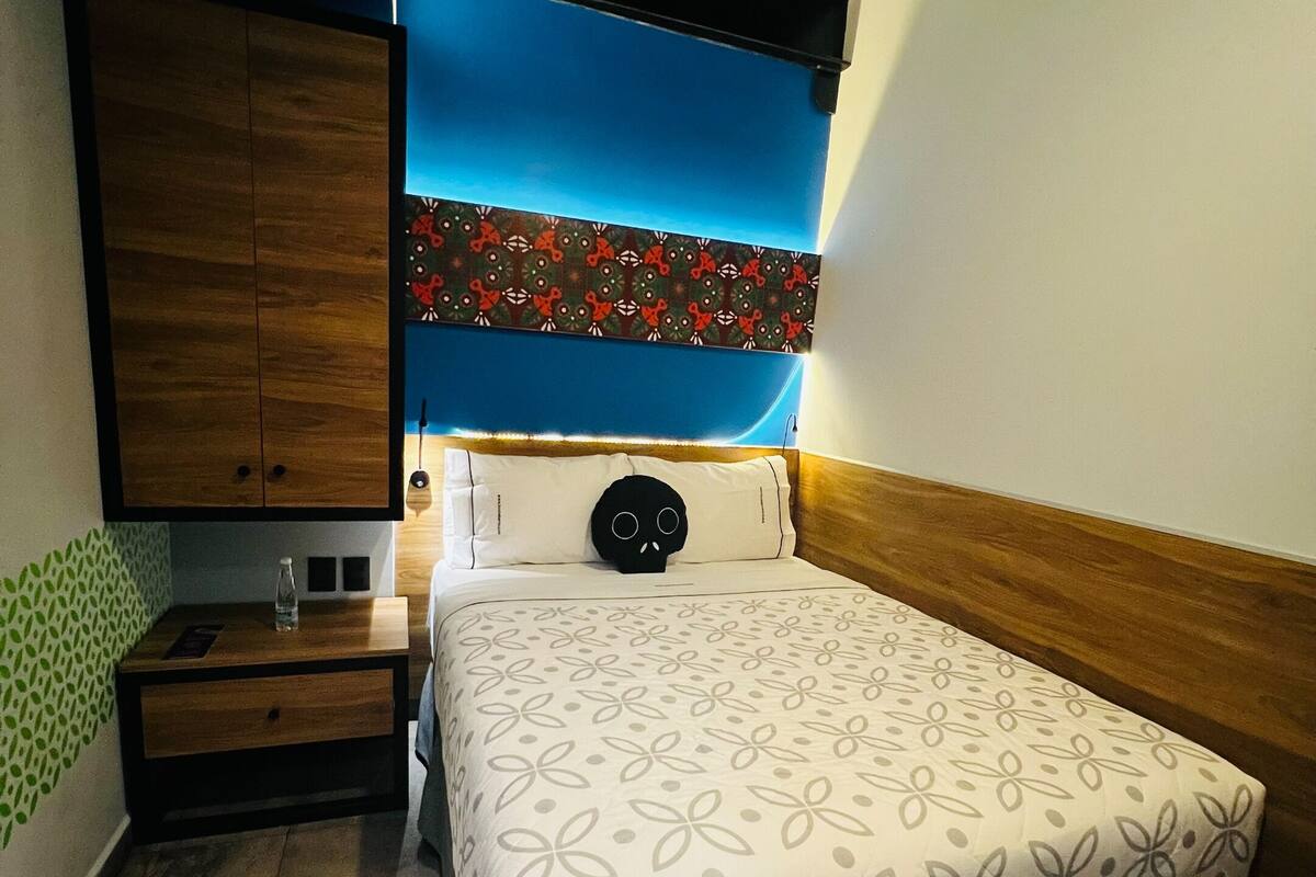 A bedroom with a bed, a nightstand, a wall-mounted shelf, and a patterned wall decoration.