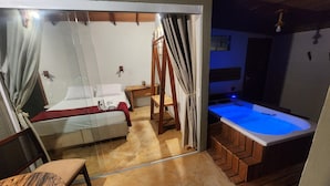 Un dormitorio con cama, un banco de madera, una mesita y un baño con bañera iluminada en azul.
