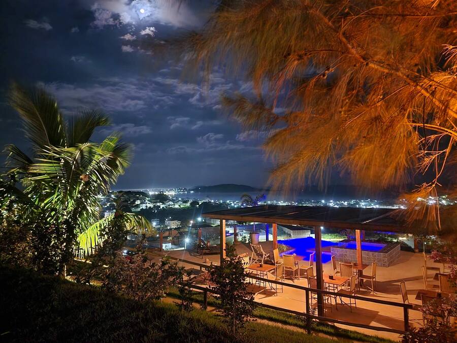 Ein Nachtblick auf ein Resort mit Außenbestuhlung und Poolbereich, umgeben von üppiger Vegetation und einer entfernten Stadtlandschaft.