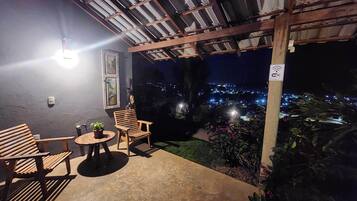 Un espacio de estar al aire libre cubierto con una mesa, sillas, una planta en maceta y vistas a un paisaje urbano nocturno.