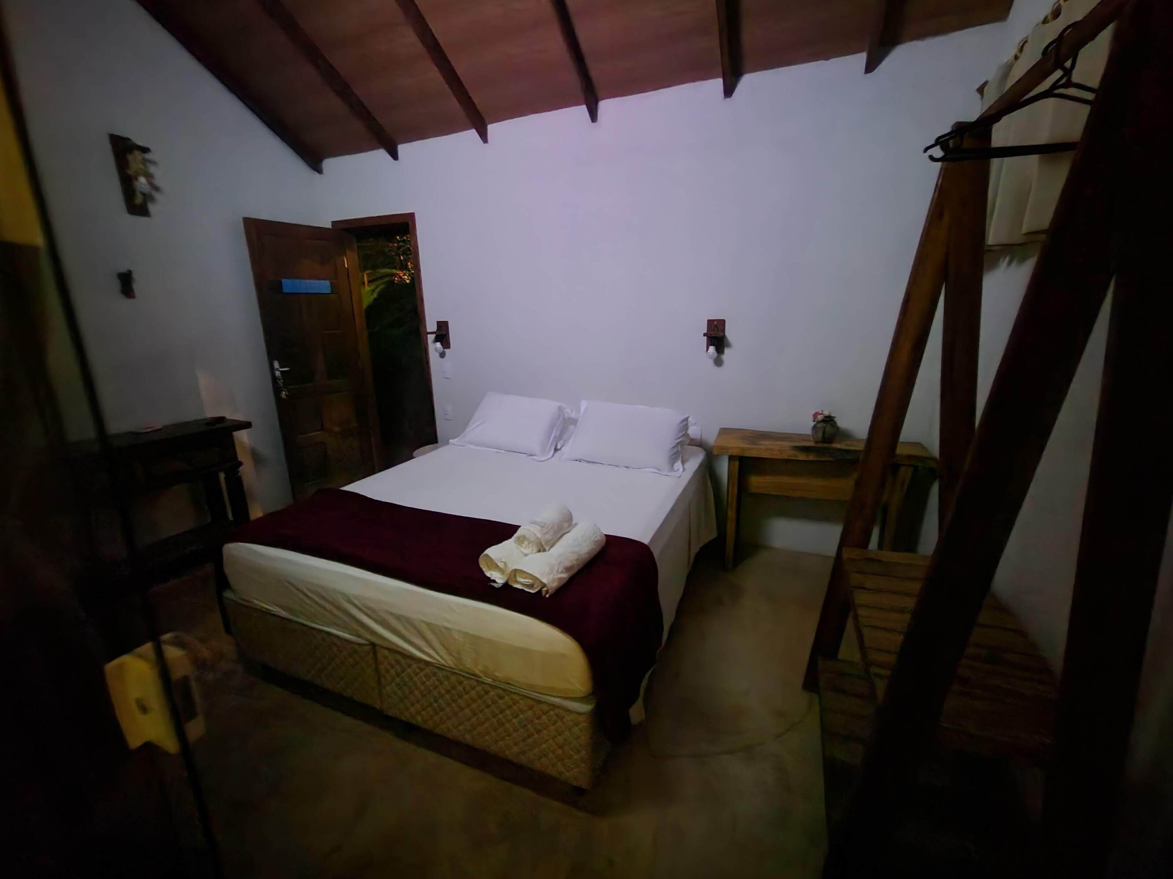 A single bed with a red blanket and white pillows, a wooden bench, a small table with a vase, and a wooden door.