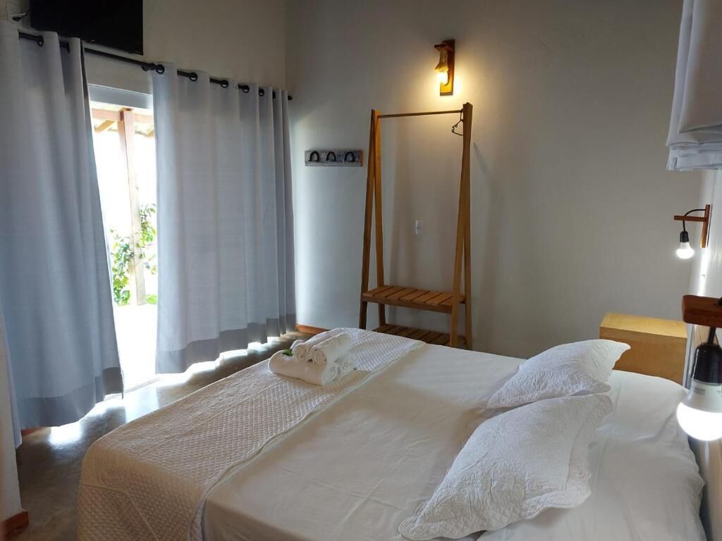 A hotel room with a bed, a wooden chair, a wall-mounted light fixture, and a window with curtains.