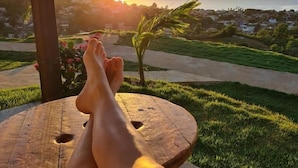 Una persona descansando con las piernas cruzadas en un banco de madera, contemplando un paisaje costero al atardecer.