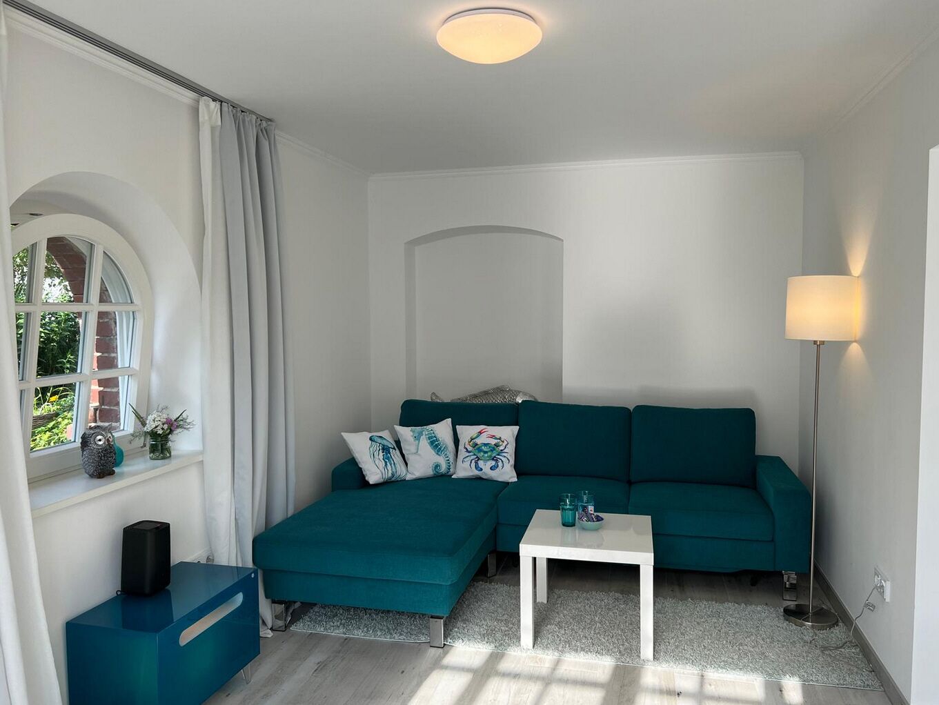 Ein modernes Wohnzimmer mit einem türkisfarbenen Sofa, einem weißen Couchtisch und einer Stehlampe. Es gibt ein Fenster mit Vorhängen, einen kleinen Tisch mit einem Lautsprecher und eine dekorative Eule.