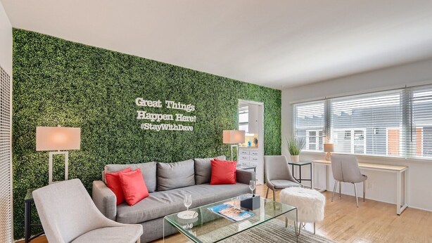 Una sala moderna con una pared verde, un sofá gris con cojines rojos, una mesa de centro de vidrio y un escritorio con dos sillas.