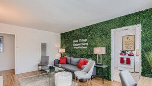 Una sala de estar moderna con una pared verde, un sofá gris, una mesa de centro de vidrio y un otoman blanco y mullido.