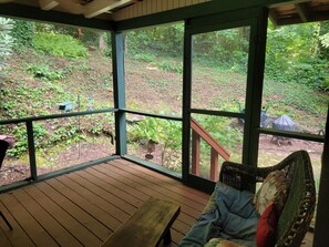 Une terrasse en bois avec un banc rembourré et un fauteuil en osier, donnant sur un jardin où poussent différentes plantes et où se trouve un barbecue.