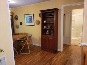 Een eetgedeelte met een houten tafel en stoelen, een hoge houten boekenkast en een lijst met een afbeelding aan de muur.