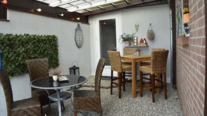 Un'area patio con sedie in vimini e un tavolo rotondo, una sveglia sul muro, e un muro in mattoni con uno scaffale che contiene bottiglie e una pianta.