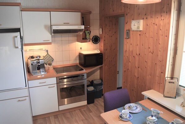 A compact kitchen with white cabinets, a built-in oven, a microwave, and a coffee machine. There is a dining area with a table set for two, featuring a blue tablecloth and white plates. - Ferienhaus Heideglück (Bad Fallingbostel)