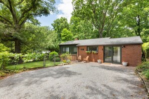 A single-story brick house with a gravel driveway, a fenced yard, and a large tree in the background.