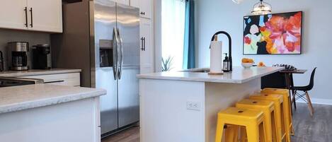 Una cocina moderna con armarios blancos, electrodomésticos de acero inoxidable y una isla central con taburetes amarillos.