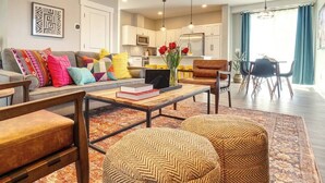 Un salon moderne avec un canapé, une table basse et des poufs. On y trouve également un coin repas avec une table et des chaises, et une cuisine en arrière-plan.