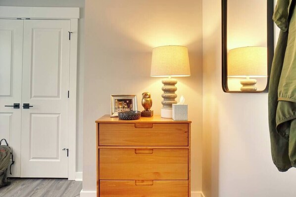 A wooden dresser with a lamp and decorative items, a white door, and a coat hanging on the wall.