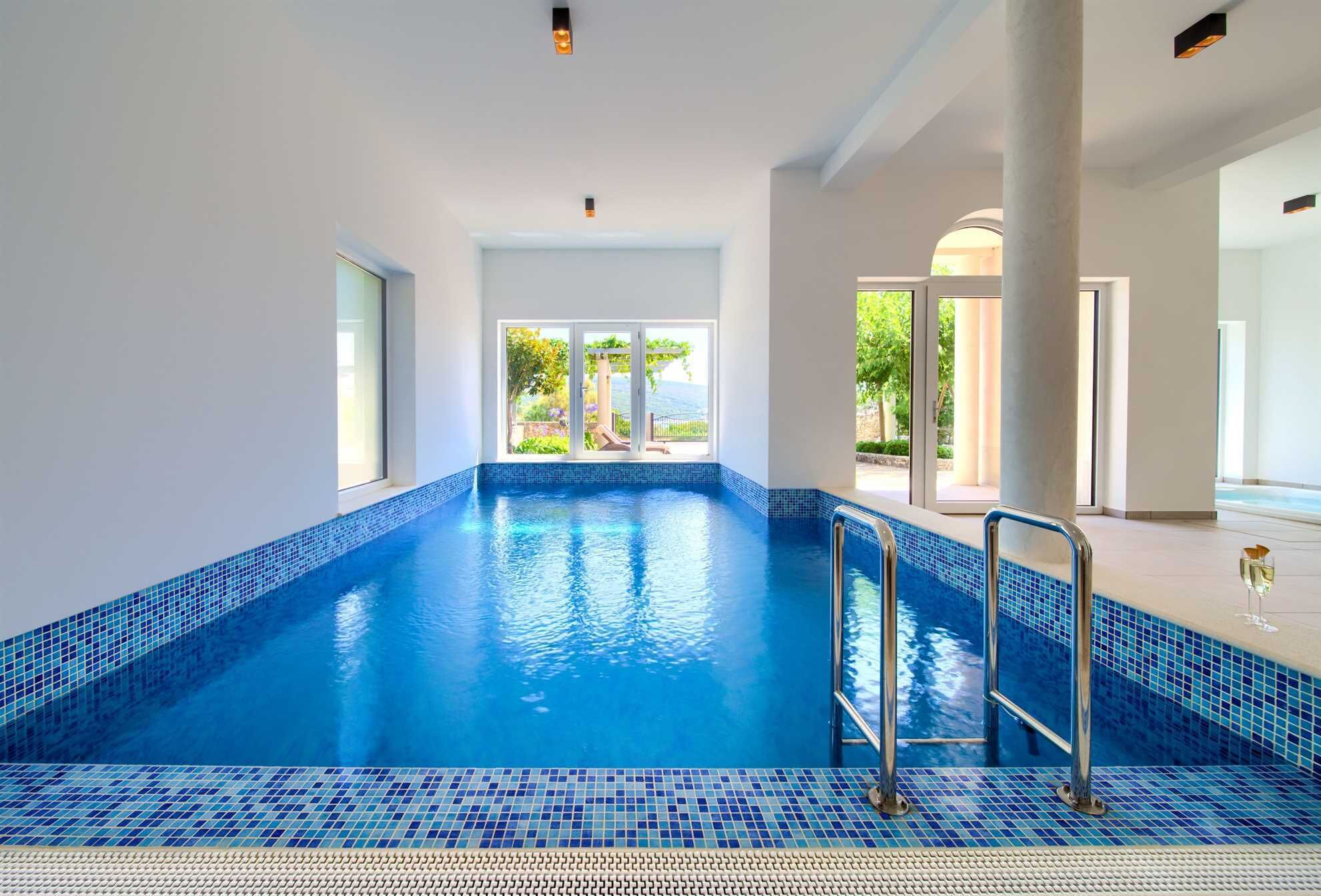 Une piscine intérieure à l’eau d’un bleu limpide, avec un sol carrelé et une vue sur l’extérieur grâce à de grandes fenêtres.