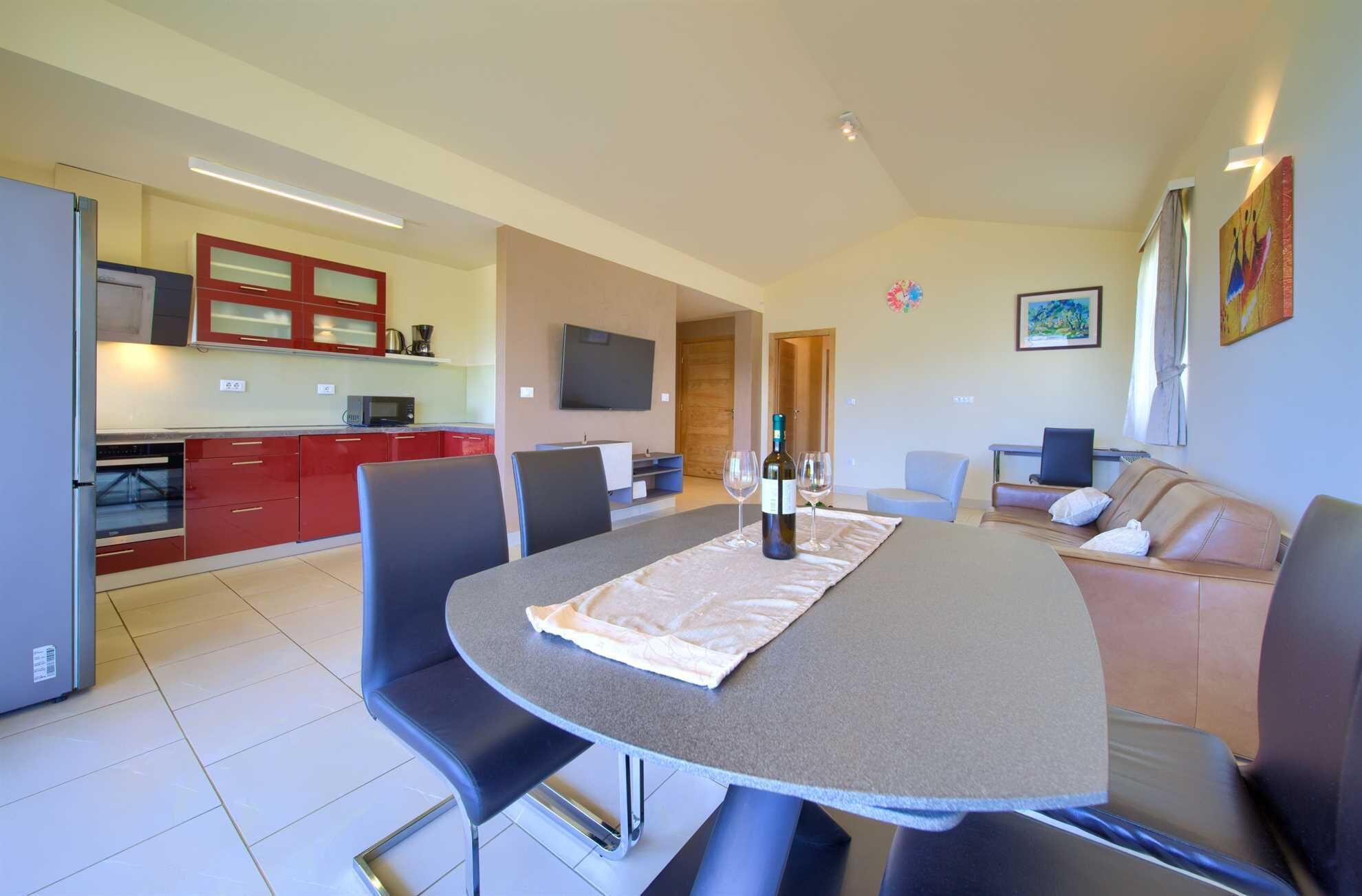 A modern kitchen with red cabinets, a dining area with a round table, and a living room with a sofa and a TV.