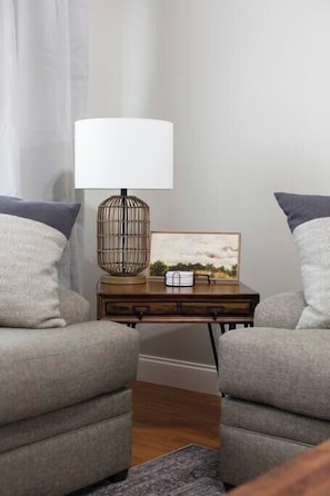 A cozy living room with two grey armchairs, a wooden side table with a lamp and picture, and a grey rug.