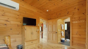 A wooden cabin interior with a television on a cabinet, a bed, and a bathroom visible through an open door. - Mountain View Cabin|Hot Tub|Fenced Yard for Pets (Mill Spring)