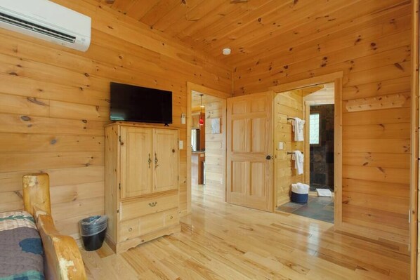 A wooden cabin interior with a television on a cabinet, a bed, and a bathroom visible through an open door. - Sunset mountain views, hot tub and fenced yard (Mill Spring)
