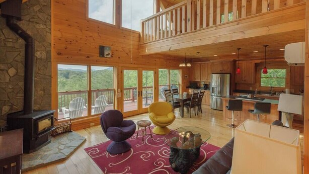 A spacious living area with a stone fireplace, a dining table, and a kitchen in the background. - Mountain View Cabin|Hot Tub|Fenced Yard for Pets (Mill Spring)
