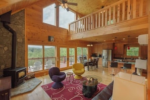 A spacious living area with a stone fireplace, a dining table, and a kitchen in the background. - Mountain View Cabin|Hot Tub|Fenced Yard for Pets (Mill Spring)