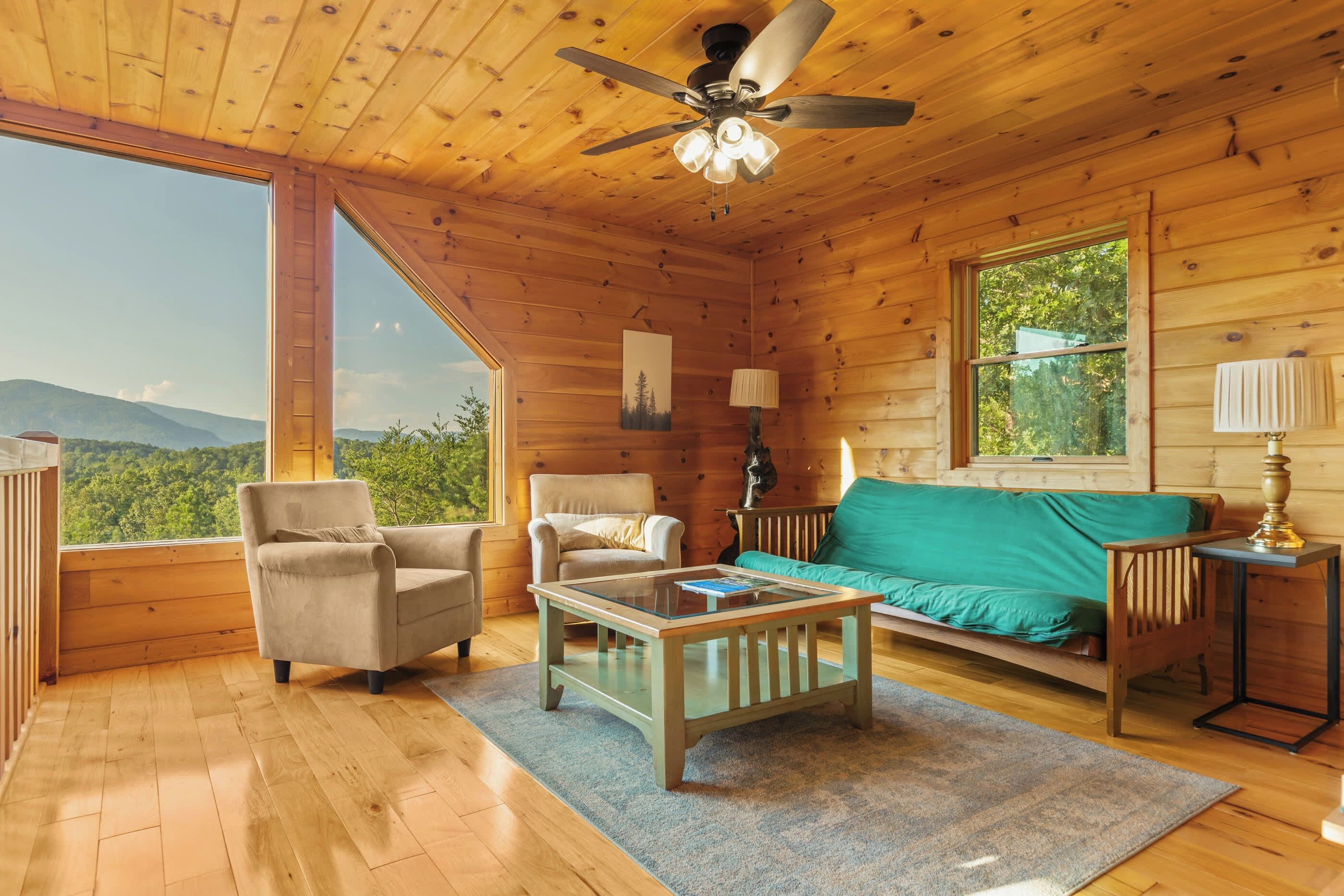 A wooden cabin interior with a ceiling fan, a sofa, two armchairs, a coffee table, and a view of the outdoors.
