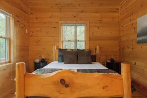 A wooden cabin bedroom with a bed, two nightstands, a window, and a painting on the wall. - Mountain View Cabin|Hot Tub|Fenced Yard for Pets (Mill Spring)
