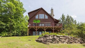 A two-story log cabin with a balcony, surrounded by a stone wall and lush greenery. - Mountain View Cabin|Hot Tub|Fenced Yard for Pets (Mill Spring)