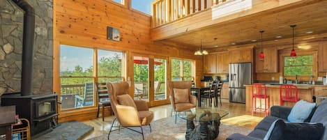 A spacious cabin interior with a stone fireplace, large windows, a dining area, and a kitchen.