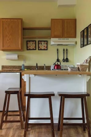 Une cuisine avec des meubles de rangement en bois, un plan de travail blanc, une bouilloire et deux tabourets.
