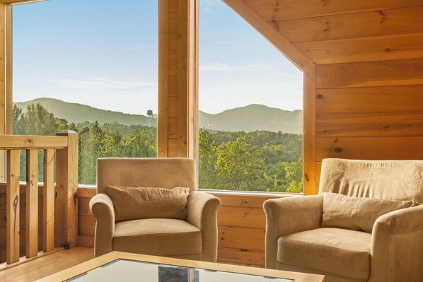 A wooden cabin interior with two armchairs and a glass-top table, overlooking a forested landscape.