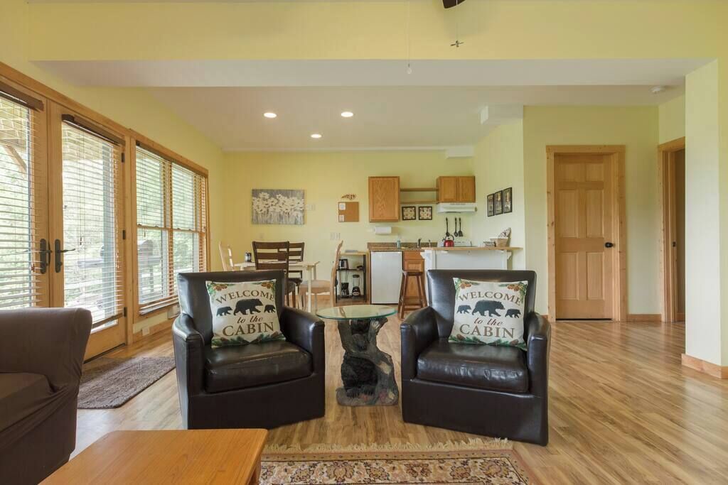 A living room with two leather armchairs, a coffee table, and a rug. There are wooden cabinets and a kitchen in the background.