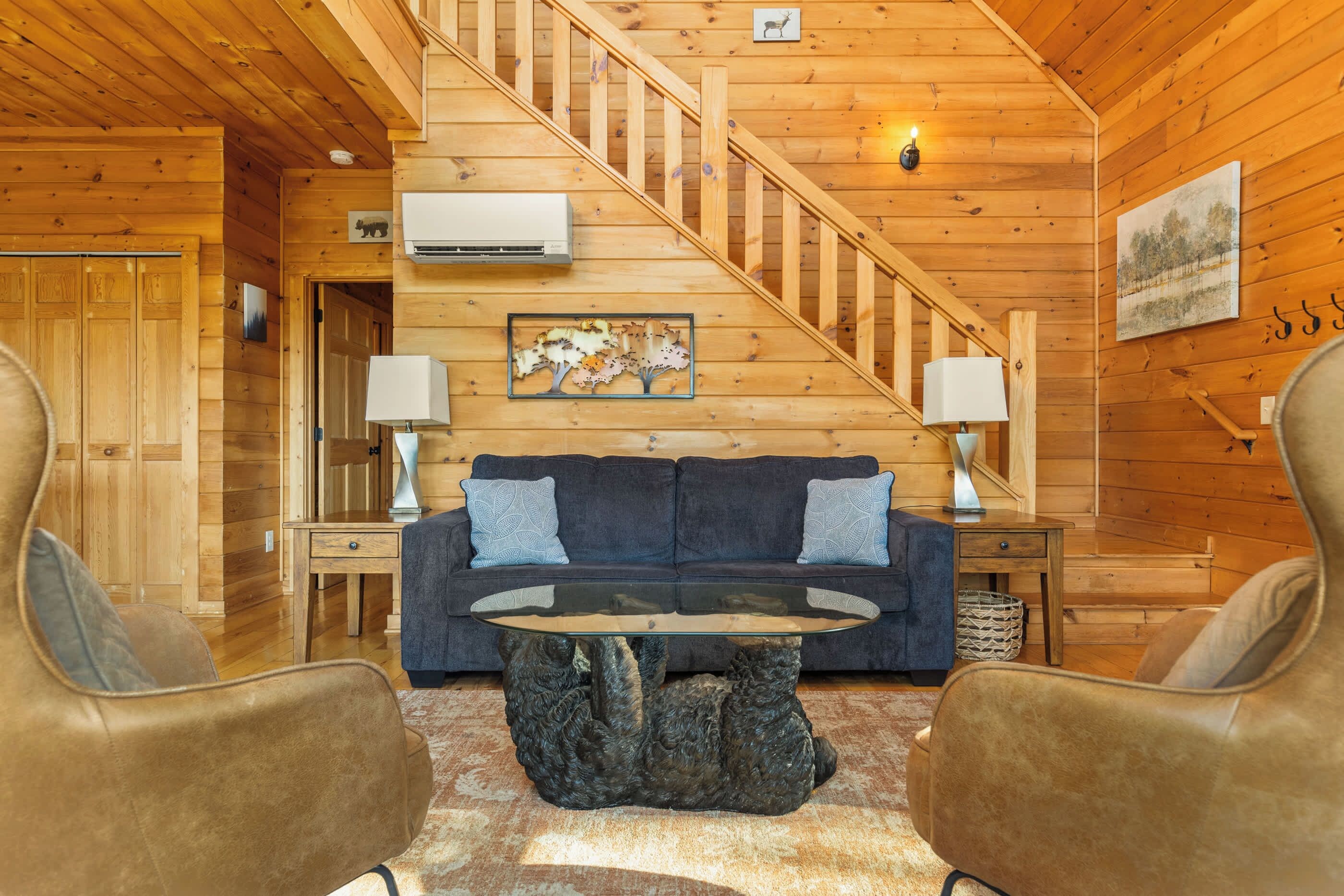 A cozy cabin interior with a wooden staircase, a sofa, two armchairs, a coffee table, and a painting on the wall.