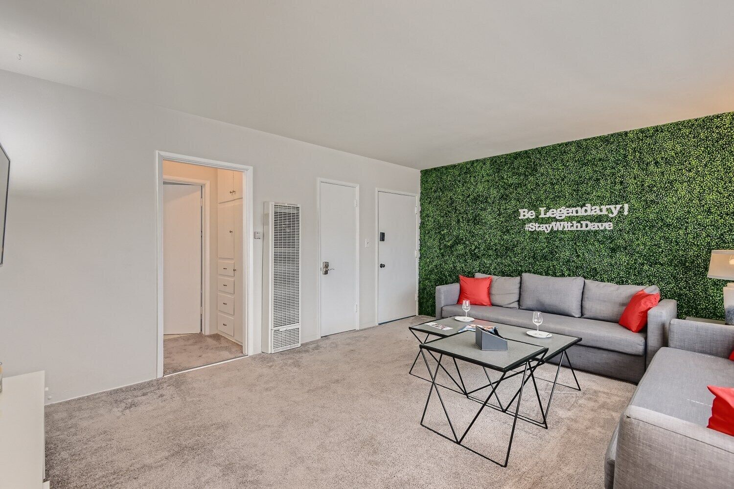 Un salon moderne avec un canapé gris, des coussins rouges et un mur vert orné d’un slogan. On y trouve une porte blanche et une grille d’aération sur le mur.