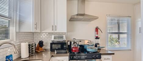 A modern kitchen with white cabinets, a stainless steel sink, a stove, a microwave, and a gas range. There is a window with blinds, a fire extinguisher, and a knife block on the counter.